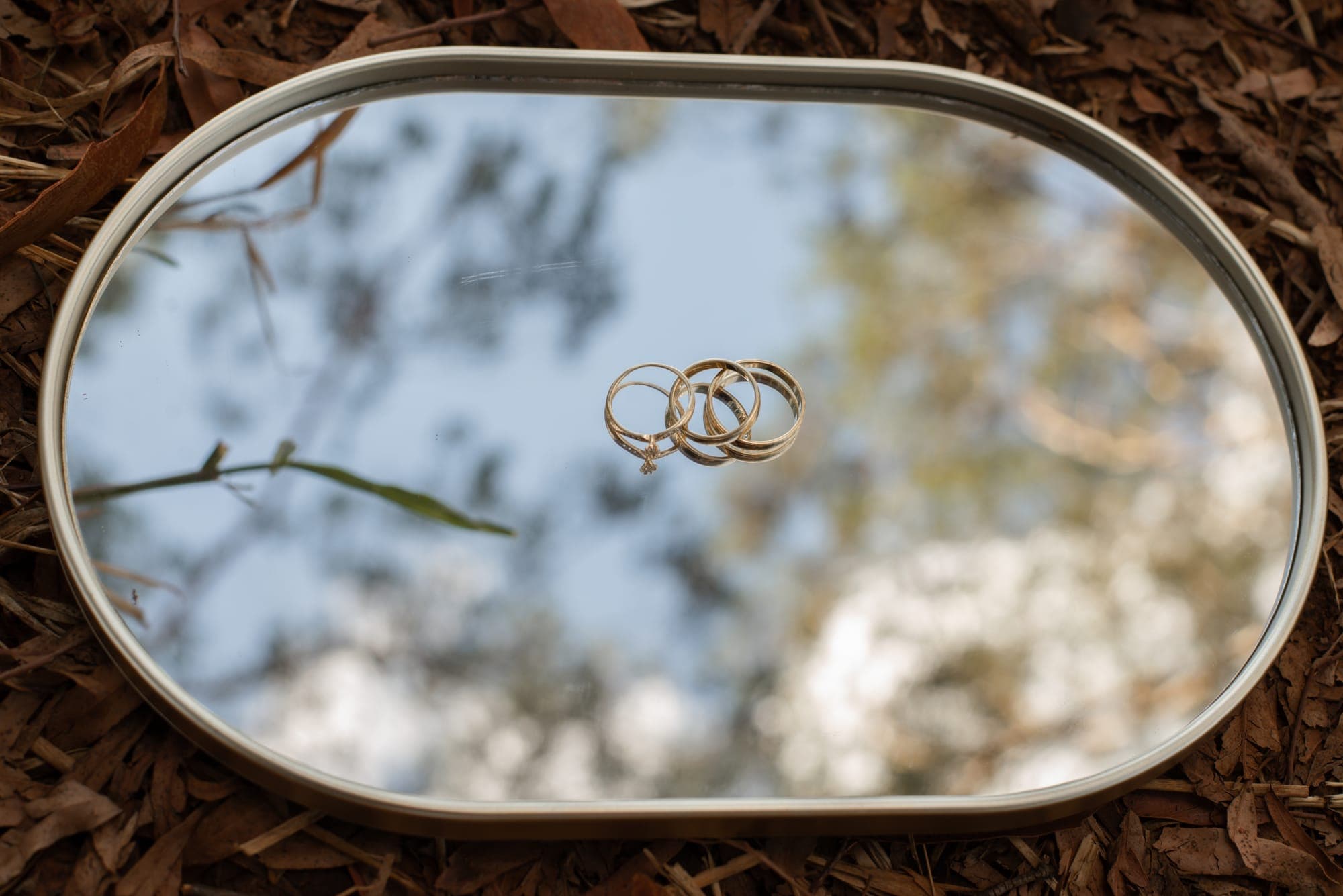 As alianças e o anel de noivado sobre um espelho oval que reflete o céu
